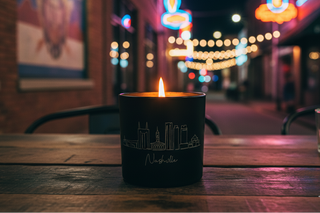 Candle in a black container with 'Nashville' engraving on a wooden table at night.