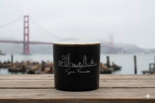 Black candle with 'San Francisco' engraving on a wooden surface with a blurred background of water and golden gate bridge.