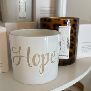 A white ceramic candle jar with the word 'Hope' in elegant gold script, displayed on a white shelf alongside leopard-print candles and white boxes.