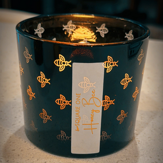 Dark glass candle with bee patterns that are illuminated and a label on a light surface