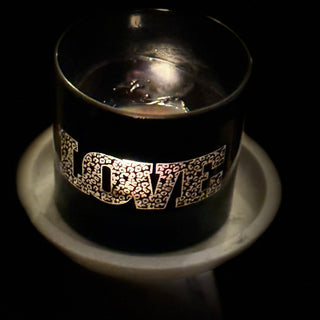 A black glass candle holder with the word 'LOVE' spelled out in sparkling silver letters, sitting on a white candlestick holder against a dark background.