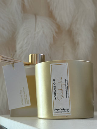 Gold candle jar with a label on a white feather background beside a scent diffuser bottle 