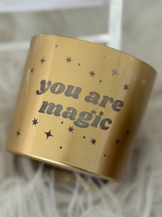 Gold candle jar with 'you are magic' text and star designs on a soft white background