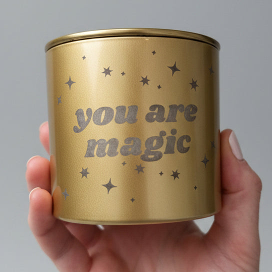 hand holding a gold 'you are magic' candle with stars and a gold lid with a white backdrop