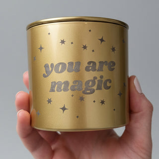 hand holding a gold 'you are magic' candle with stars and a gold lid with a white backdrop