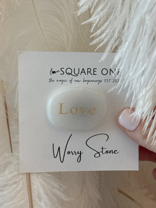 White stone with 'Love' engraved, held on a card with 'Wavy Stone' and branding text, against a soft feather background.