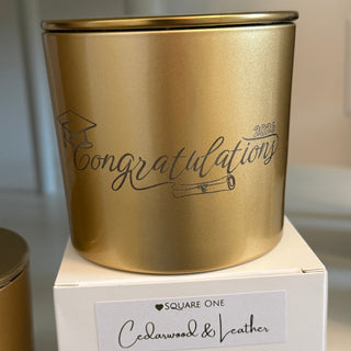 A gold metal candle tin next to its white packaging box. The tin features the word 'Congratulations' printed in black script, and the box has a white label reading 'Square One Candlewax & Leather'.