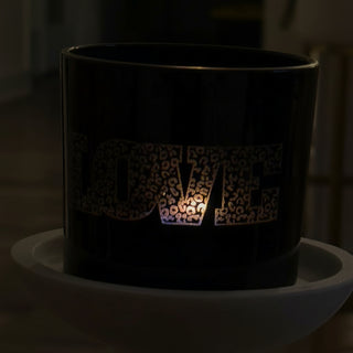 A black cylindrical candle holder or tumbler with the word "LOVE" cut out in a leopard-print pattern, glowing with warm light from within, displayed on a white marble pedestal in an indoor setting.