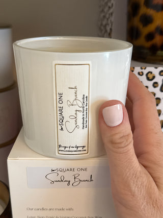 Hand holding a white candle with a label on a neutral background