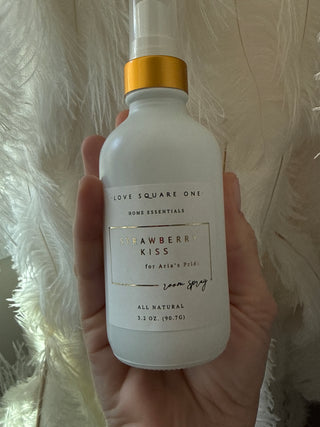 Hand holding a strawberry kiss room spray bottle with a gold cap on a textured white background