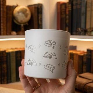 A hand holding a white engraved candle tin with a gold-lid and fine line-art illustrations of books and stars.