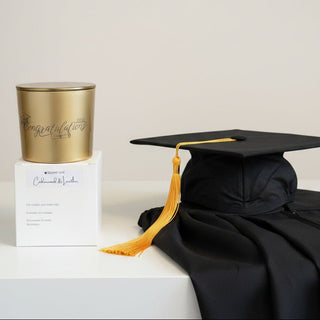 A gold metal candle tin next to its white packaging box. The tin features the word 'Congratulations' printed in black script, and the box has a white label reading 'Square One Candlewax & Leather'.