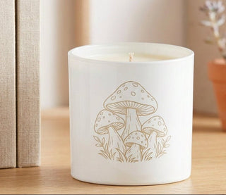 A white cylindrical jar candle with a metallic gold lid and a delicate line-art illustration of mushrooms on the front, resting on a light-colored surface.