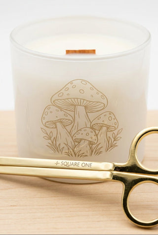 A white glass candle jar with a mushroom line-art design and a small wooden wick, shown on a light wood surface with gold scissors in the foreground.
