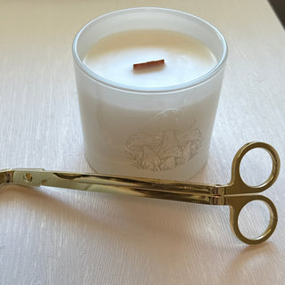 A white wax candle in a clear glass jar with a wooden wick, placed on a light wood table next to a pair of gold metal scissor tongs. In the background, another candle is visible on a shelf.