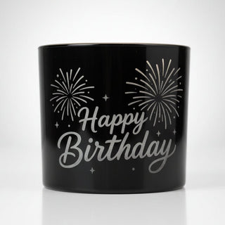 Glossy black ceramic Happy Birthday mug with white script and fireworks illustrations on a speckled countertop.