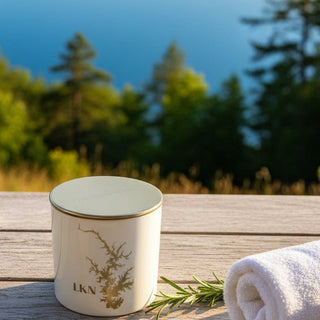A white glass candle jar with a metallic gold lid, featuring an etched lake design and the initials 'LKN', resting on a wooden dock. Au generated image. Etching is clear not gold 