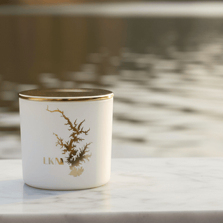 White cylindrical candle jar with a gold lid and gold dendritic tree design on the front, resting on white faux fur.
