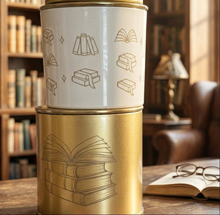 A close-up of a white glass candle jar  with a gold metallic top and base, decorated with simple line drawings of books and open book motif wrap