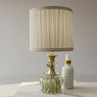 A white bottle with a gold spray nozzle and a clear ribbed glass decanter with a gold-toned cap, arranged on a white marble surface next to a pleated beige lampshade against a dark gray wall.