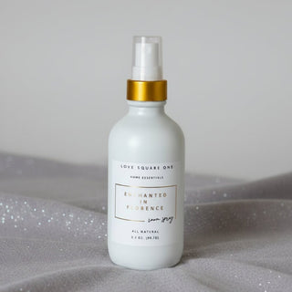 A white room spray bottle of Love Square One Enchanted in Florence All Natural Room Spray, featuring a gold collar and clear spray nozzle, resting on a grey surface.