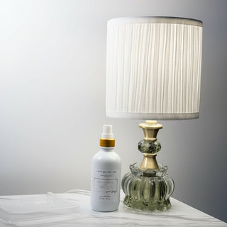 A white room spray bottle with a gold collar and clear cap, placed next to a ribbed white lampshade and a green glass decorative object on a marble-like surface.