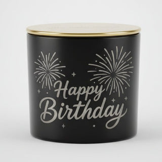 A black cylindrical candle tin with a gold lid, featuring a 'Happy Birthday' design with white script and fireworks.