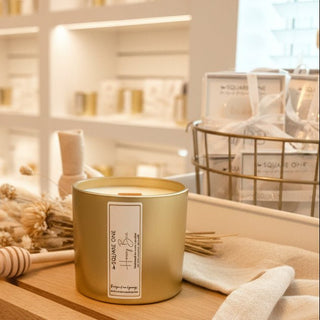 Large gold Square One Honey Bee candle jar on a shelf in a bright retail display near a window.