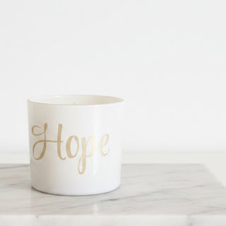 A white ceramic candle jar with the word 'Hope' in elegant gold script, displayed on a white shelf alongside leopard-print candles and white boxes.