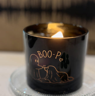 A black glass jar candle with a lit flame, featuring a gold foil illustration of a ghost and the text 'BOO-P' on the front, set on a clear glass coaster.
