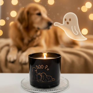 A black glass jar candle with a lit flame, featuring a gold foil illustration of a ghost and the text 'BOO-P' on the front, set on a clear glass coaster.