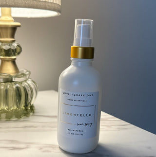 A white room spray bottle of Love Square One Limoncello home essentials, featuring a gold collar and clear spray top, set on a marble surface next to a lit lamp.