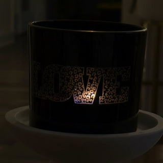 A black cylindrical candle holder or tumbler with the word "LOVE" cut out in a leopard-print pattern, glowing.