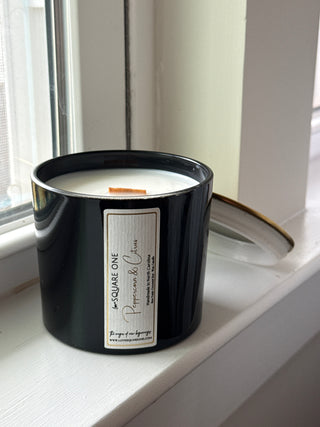 A black love square one candle jar with a white label reading 'Love SQUARE ONE' and 'Peppercorn & Citrus', photographed leaning against a white window frame.