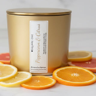 A gold metal candle tin lid in a warm gold finish with a white label reading 'Love Square One Peppercorn & Citrus,' styled with AI sliced citrus fruits on a light surface.