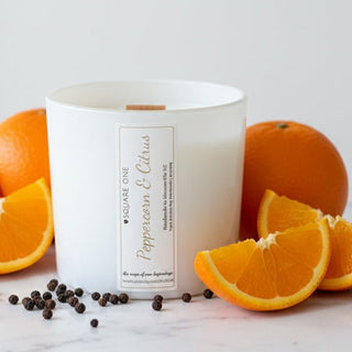 A white glass candle jar labeled 'Love Square One Peppercorn & Citrus' surrounded by AI generated fresh orange slices, whole oranges, and scattered black peppercorns on a light marble surface against a white background.