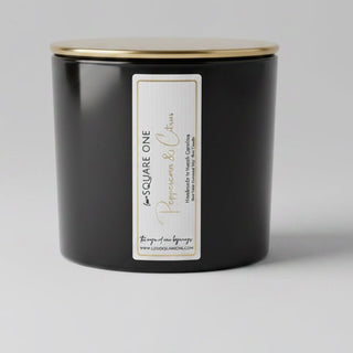 A black love square jar candle with a gold-toned rim and a white label reading ' Love SQUARE ONE' and 'Peppercorn & Citrus', photographed in a white studio interior setting.