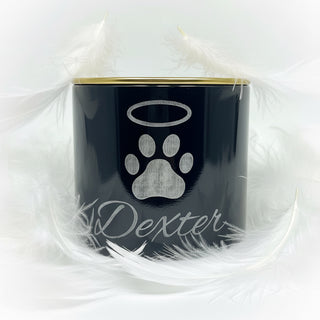 A black cylindrical keepsake candle jar with a gold lid and a etched paw print and cursive name design, placed on a white textured blanket next to a handwritten note.