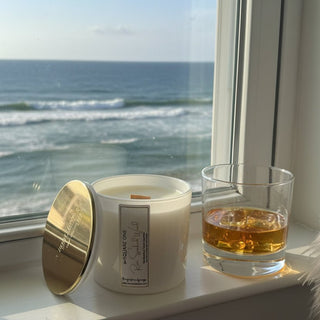 A white candle jar with a gold lid leaning beside it on a windowsill, next to a glass of amber liquor, with the ocean visible outside. ai ocean and drink.