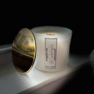 A white candle jar with a gold-toned lid leaning against it on a windowsill, lit from the side against a dark background. The front label reads love square one rum soaked wood'.