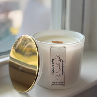A white glass jar candle with a gold lid resting beside it on a windowsill. The front label reads 'SQUARE ONE' and 'Rose Sandalwood'.