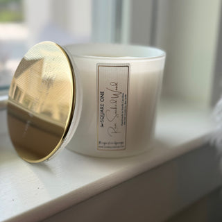 A white candle jar with a gold lid resting on a white windowsill, with a soft-focus background of a window and outdoor scenery.