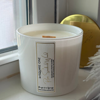 A large white jar candle with a rectangular cream label reading 'SQUARE ONE' and 'Rose Scented Wood' sits on a windowsill. Natural light from the window highlights the left side of the jar, with a gold decorative object and white feathers visible in the background.