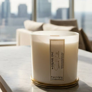 Close up of a white Square One Spice and Vanilla candle jar with a gold rim and front label, with candles blurred behind it.
