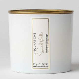 Close up of a SQUARE ONE Spice Vanilla candle tin by The Artisan Candle Company, stacked on a white shelf with other candles.