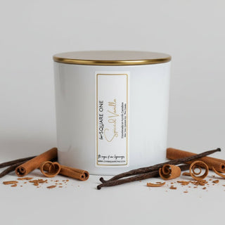 Close up of a white SQUARE ONE Special Vanilla jar candle by The Shop of Our Legacy, with other candles on a shelf.
